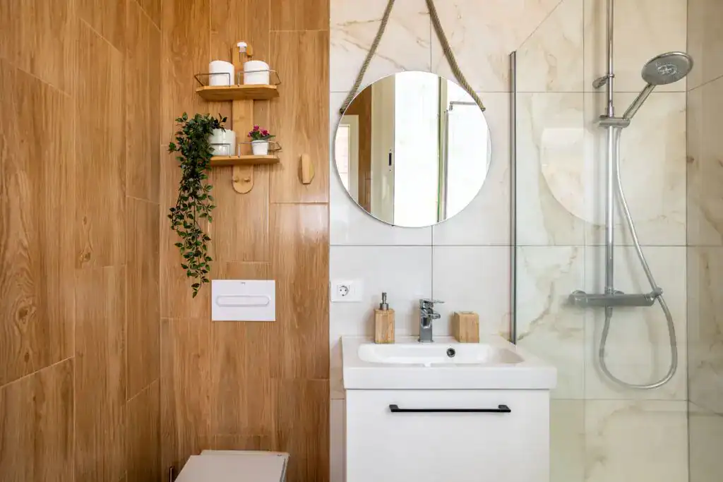 A modern bathroom with a wood-look tile feature wall, a white vanity with a round rope mirror, and a walk-in shower with marble tile.