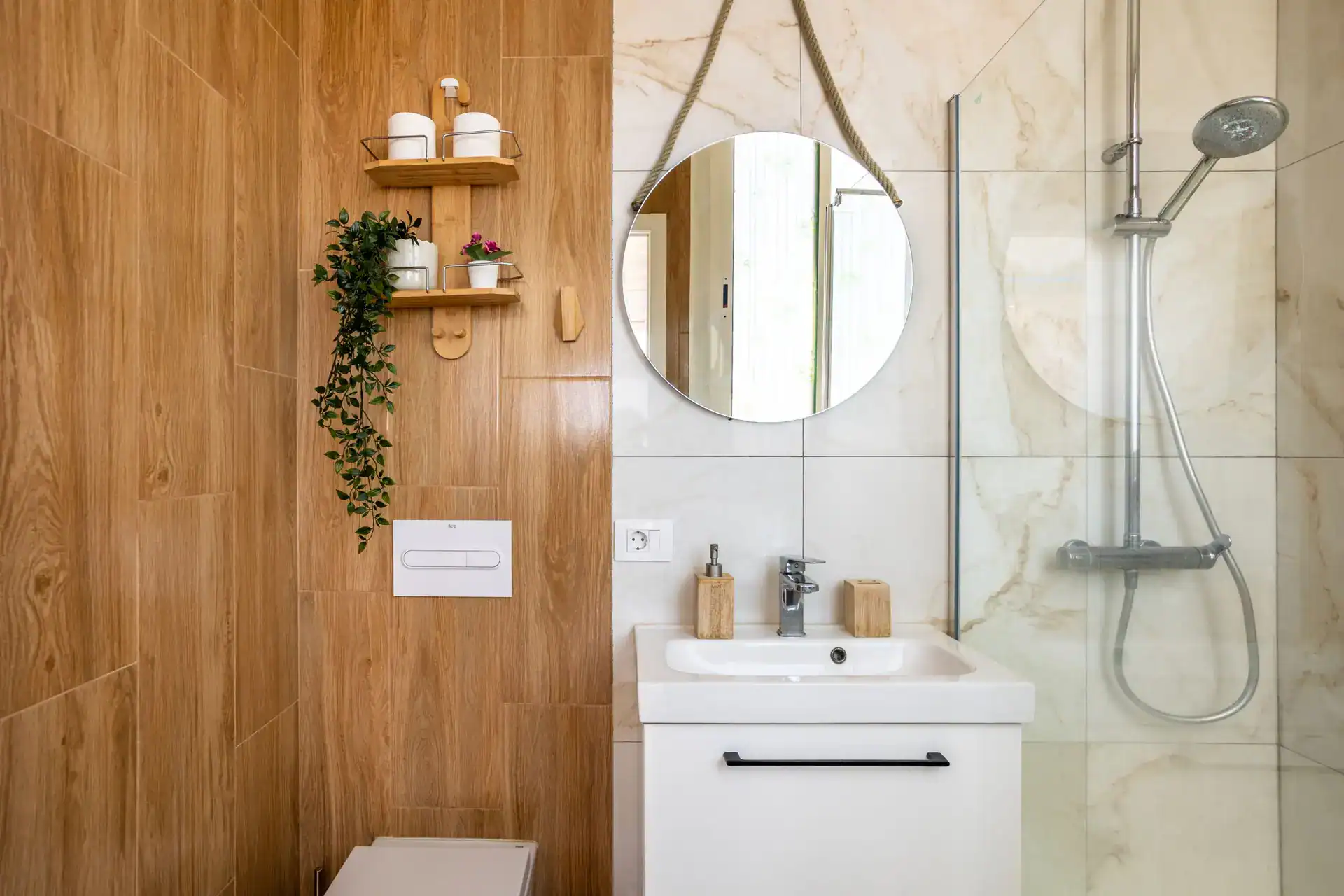 A modern bathroom with a wood-look tile feature wall, a white vanity with a round rope mirror, and a walk-in shower with marble tile.